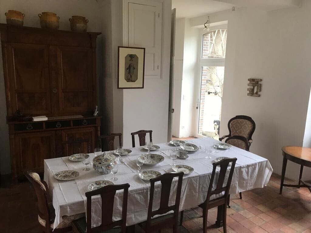 En matplats med ett bord dukat för fyra personer, omgiven av trästolar. Ett träskåp med dekorativa föremål står till vänster.