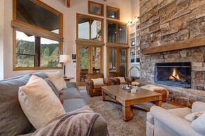 A cozy living room with a fireplace, large windows, and a stone wall. - Abode on Success View, ski-in/ski-out, hot tub, pool table, Deer Valley views (Park City)