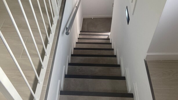 Una escalera con barandales blancos y un pasamanos de madera que lleva a un rellano con un interruptor de luz y un detector de humo.