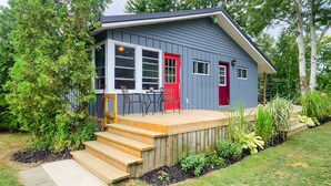 A small house with a deck, a red door, and a window overlooking a garden.