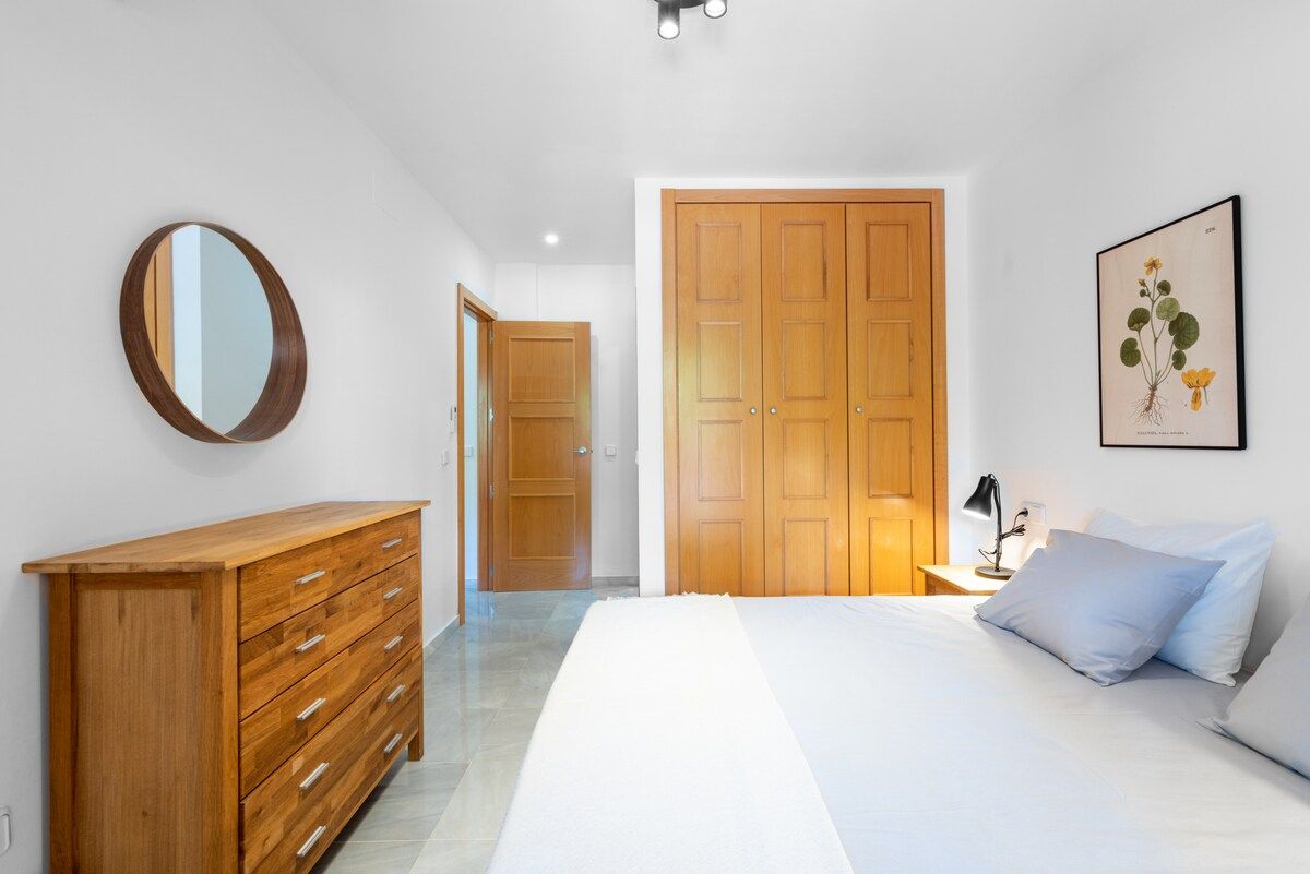 A bedroom with a wooden dresser, a large bed, a nightstand with a lamp, and a framed botanical illustration on the wall.