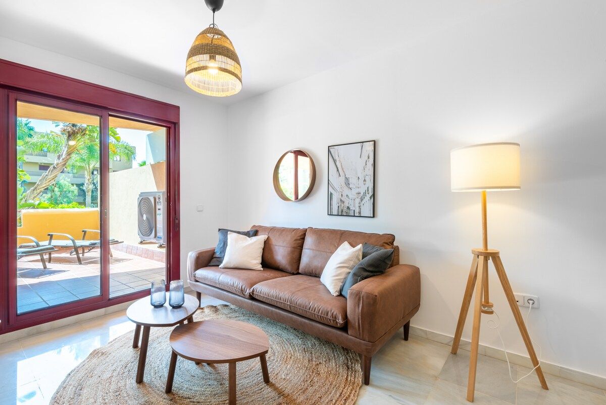 A living room with a brown leather sofa, a round wooden coffee table, a floor lamp, and a large window with a view of outdoor furniture.