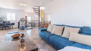 A living room with a blue sofa, a glass coffee table, a spiral staircase, and a dining area with a piano. - El Arco Iris de la Montana Sayalonga by Ruralidays (Málaga)