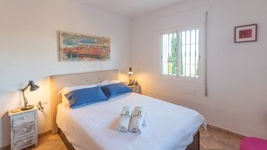 A neatly made bed with blue pillows, a bedside lamp, and a painting above it. - El Arco Iris de la Montana Sayalonga by Ruralidays (Málaga)