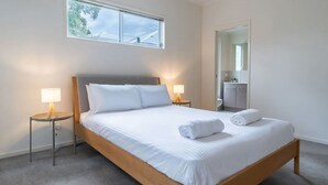 Ein ordentlich bezogenes Bett mit weißen Leinen und zwei Kissen, begleitet von Nachttischen mit Lampen, in einem Zimmer mit einem großen Spiegel und einem Fenster mit Blick auf Bäume.