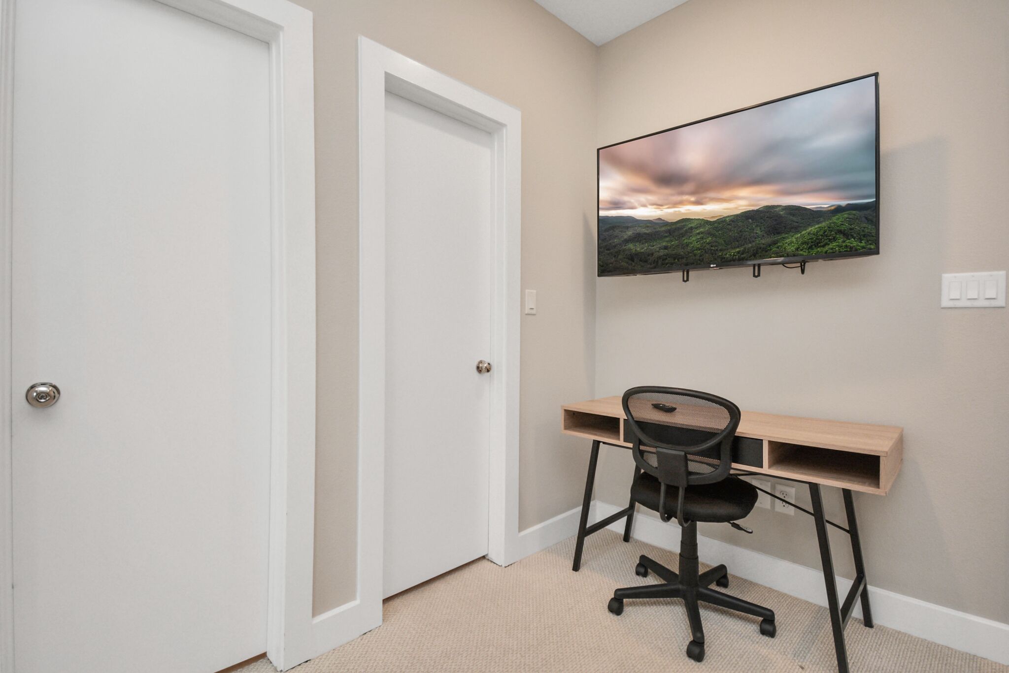 Ein Zimmer mit beigefarbenem Teppich, einem Holztisch mit schwarzem Bürostuhl, einem an der Wand befestigten Flachbildfernseher und einer weißen Tür.