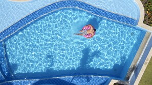 Una piscina con agua azul cristalina, rodeada por un solado de baldosas y una zona ajardinada.