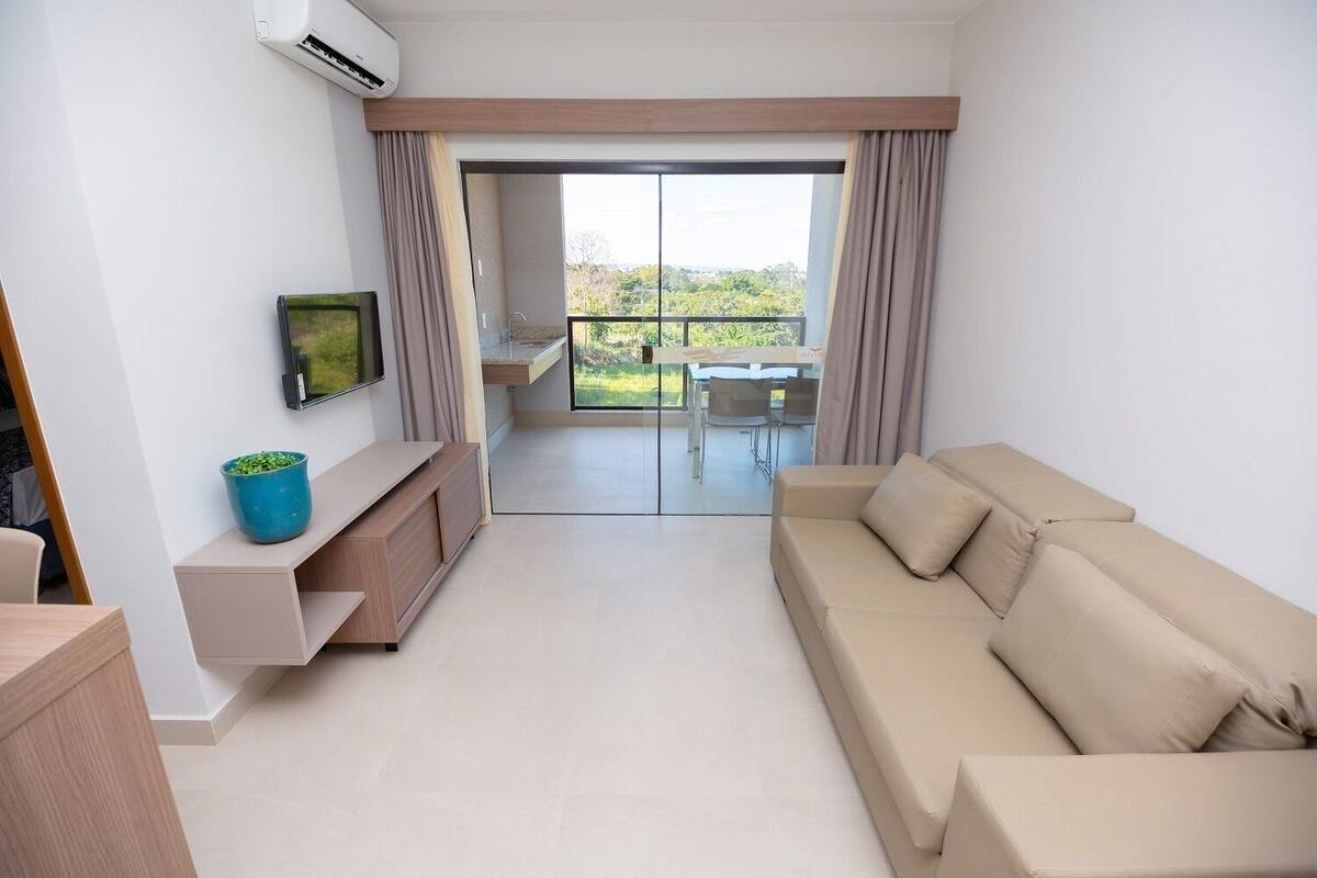 Una sala de estar moderna con un sofá color beige, un televisor montado en la pared, y un balcón con una mesa y sillas de comedor.