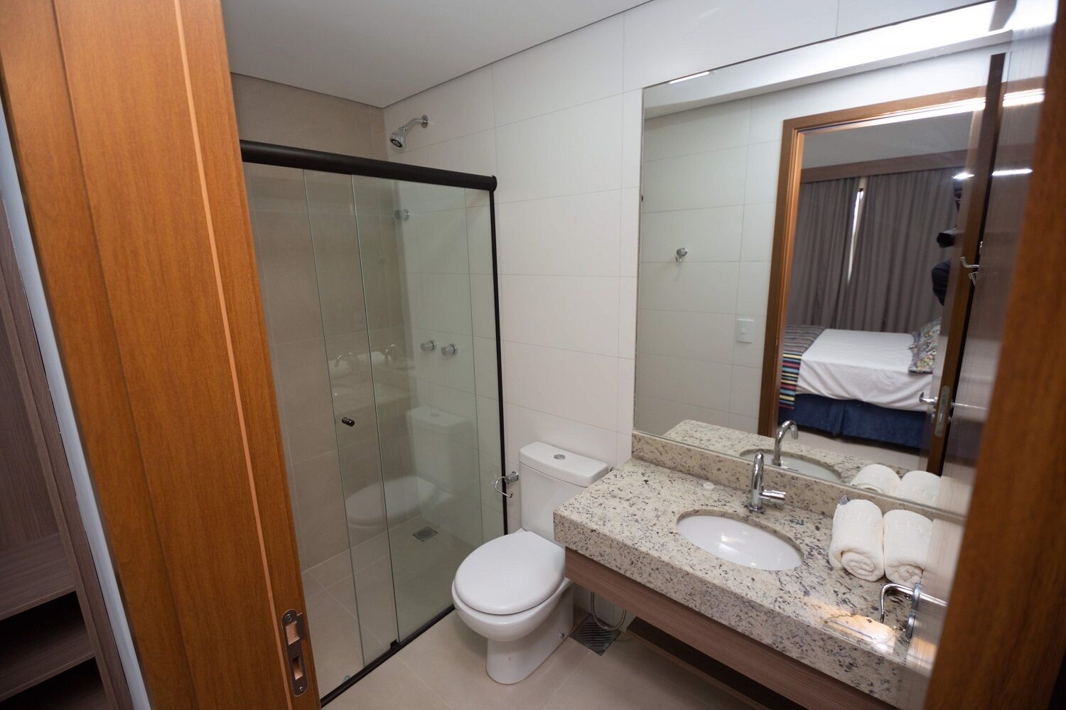 A modern bathroom with a glass-enclosed shower, a white toilet, a granite countertop sink, and a large mirror reflecting a bed.