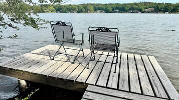 Un muelle de madera con dos sillas metálicas mirando a un lago tranquilo, rodeado de árboles y casas a lo lejos.