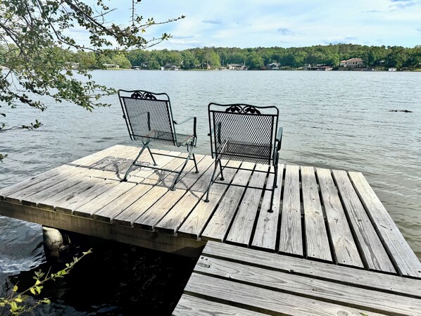 Une jetée en bois avec deux chaises en métal, donnant sur un lac paisible, entourée d’arbres et de maisons au loin.