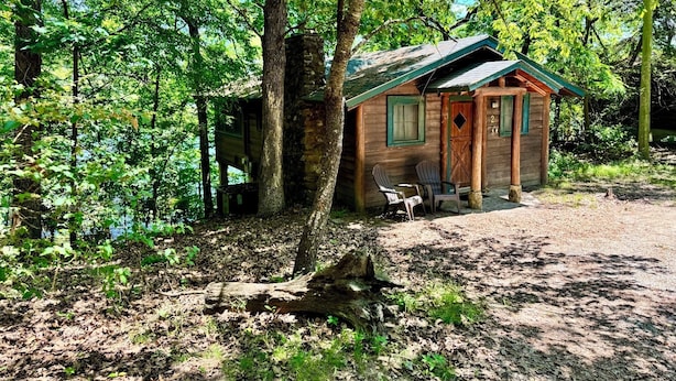 A rustic cabin with a green roof, wooden walls, and a small porch with two Adirondack chairs.