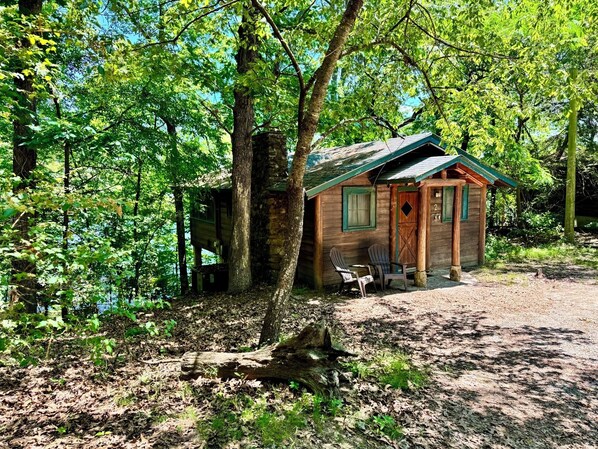 A rustic cabin with a green roof, wooden walls, and a small porch with two Adirondack chairs.