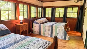 Ein Zimmer mit zwei Betten, Holzboden, einem Nachttisch mit Lampe und großen Fenstern mit Vorhängen.