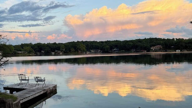 A calm lake with a wooden dock and chairs, surrounded by trees and houses.