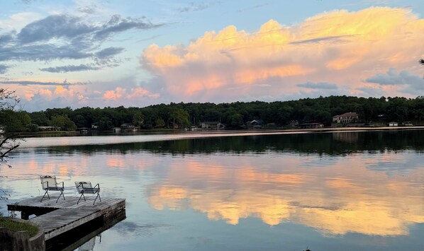 A calm lake with a wooden dock and chairs, surrounded by trees and houses.