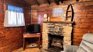 A cozy cabin interior with a stone fireplace, wooden walls, a television on a wooden stand, and a window with curtains.