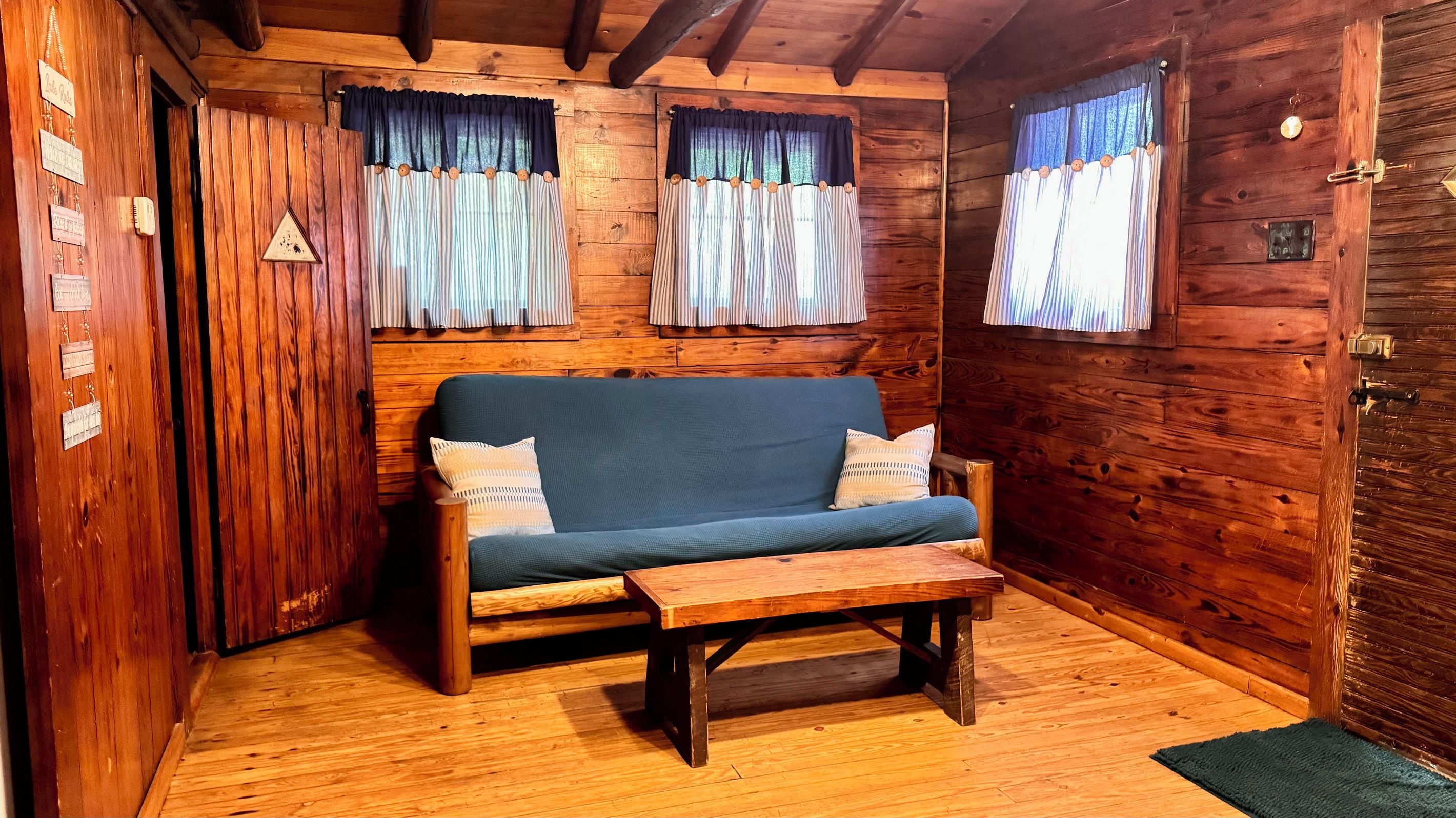 Interior de una cabaña de madera con un sofá, una mesita y cortinas en las ventanas.