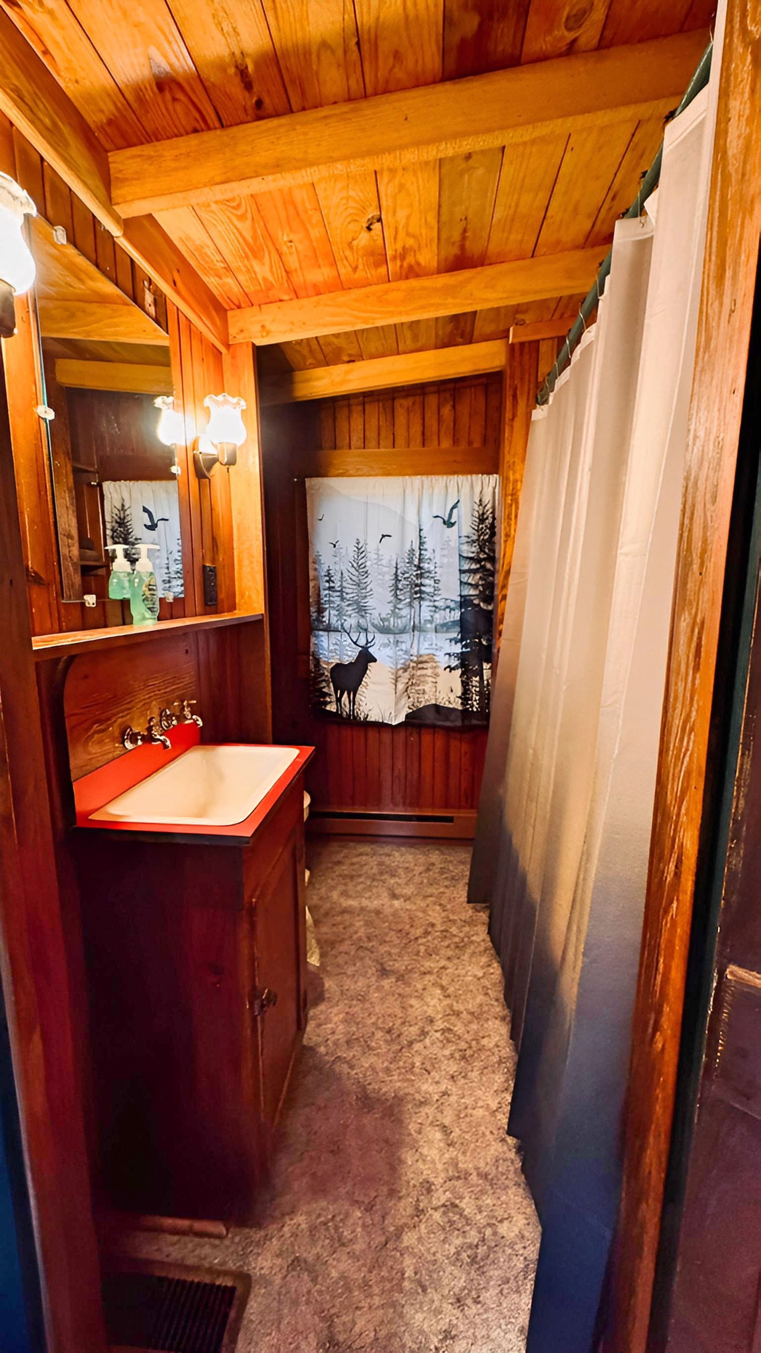 A wooden cabin interior with a wooden ceiling, a sink with a red countertop, a mirror with lights, and a window with a forest view.