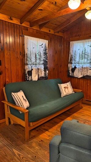 A wooden cabin interior with a green sofa, two pillows, and a window with a forest scene.