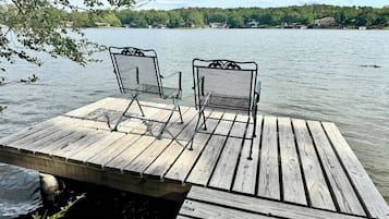A wooden dock with two metal chairs facing a calm lake, surrounded by trees and houses in the distance.