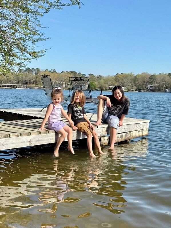 Three people sitting on a wooden dock by a lake with chairs and a clear sky in the background.
