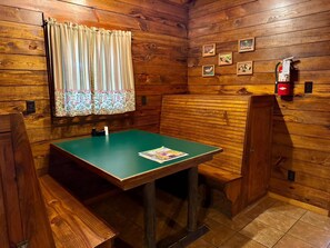 Une cabane en bois avec une table verte, des bancs, une fenêtre avec des rideaux et des tableaux encadrés accrochés au mur.