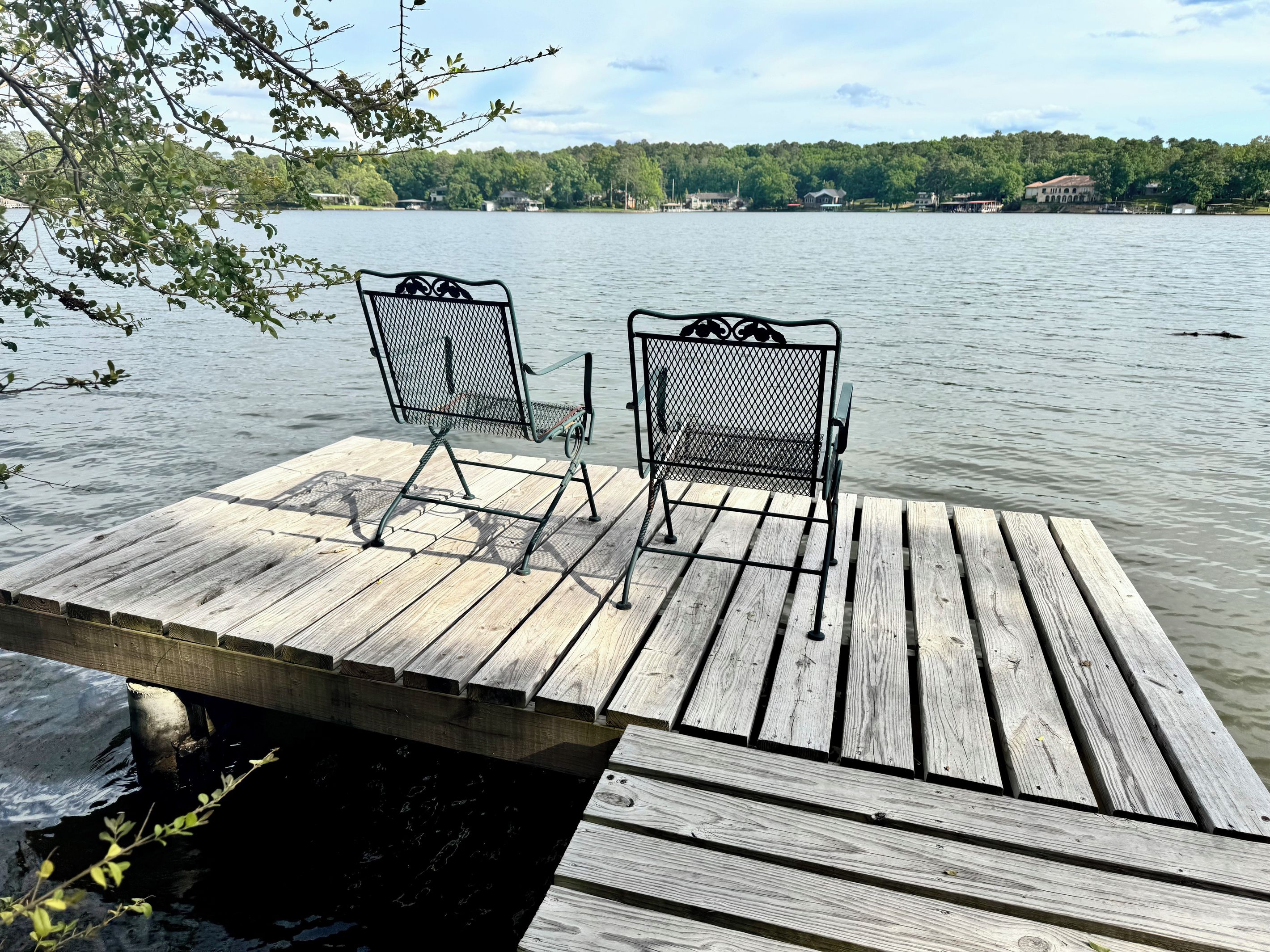 A wooden dock with two metal chairs facing a calm lake, surrounded by trees and houses in the distance.