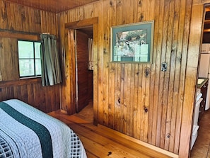 A wooden cabin room with a bed, a framed picture, and a door leading to another room.