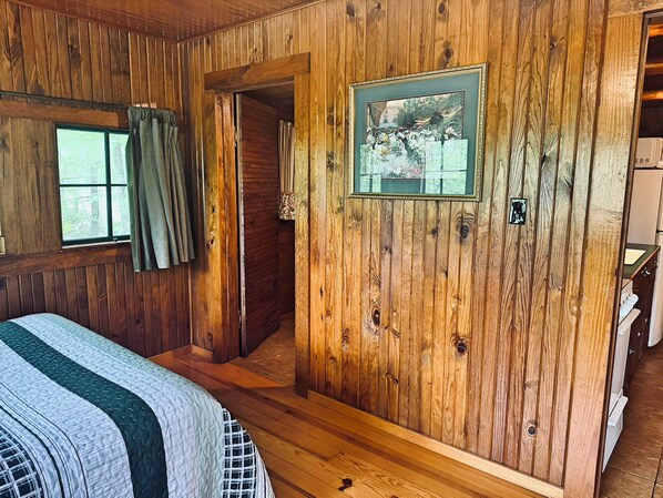 Una habitación de cabaña de madera con una cama, un cuadro enmarco y una puerta que da a otra habitación.
