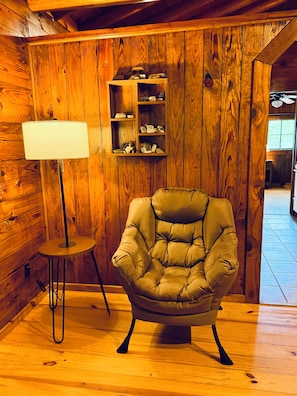 A cozy room with wooden walls, a cushioned armchair, a small table with a lamp, and a shelf with decorative items.