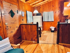 A wooden interior with a wooden door, a table, a chair, a bench, a trash bin, and a hanging light fixture.