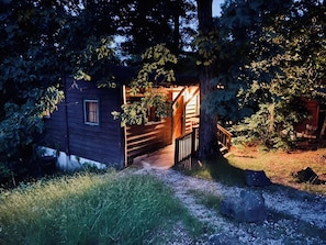 A wooden cabin with a wooden deck and a window, surrounded by trees and grass.