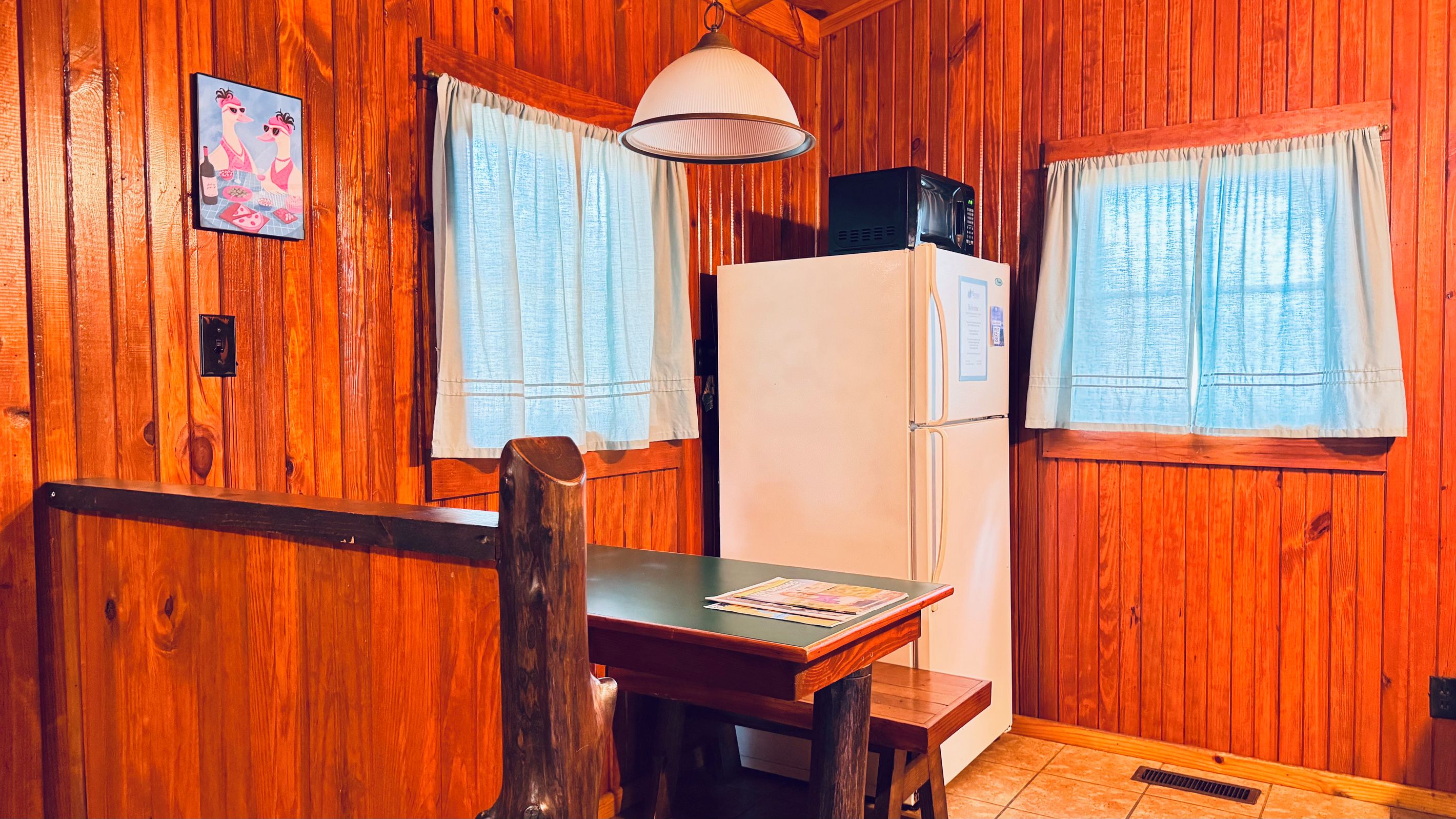 A wooden interior with a dining table, chairs, a refrigerator, and a hanging lamp.