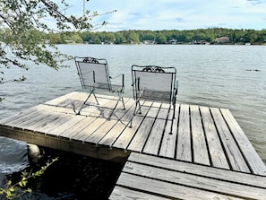 A wooden dock with two metal chairs facing a calm lake, surrounded by trees and houses in the distance.
