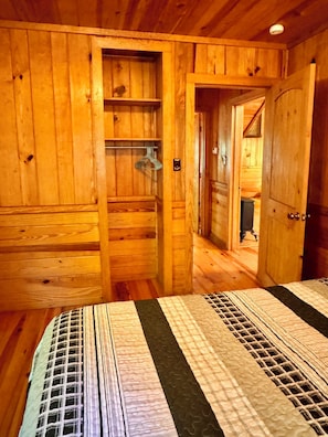 L’intérieur d’une cabane en bois, avec un lit, un portant à vêtements suspendu et une porte donnant sur une autre pièce.