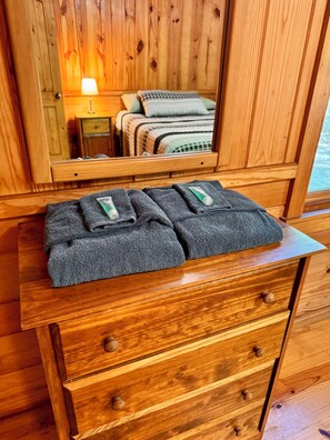 L’intérieur d’un chalet en bois, avec une commode en bois, un miroir reflétant un lit recouvert d’une couverture verte, et une table de chevet avec une lampe.