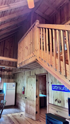 Un intérieur en bois avec un escalier menant à un niveau supérieur, un réfrigérateur et une pancarte sur laquelle on peut lire : « À bientôt ! »