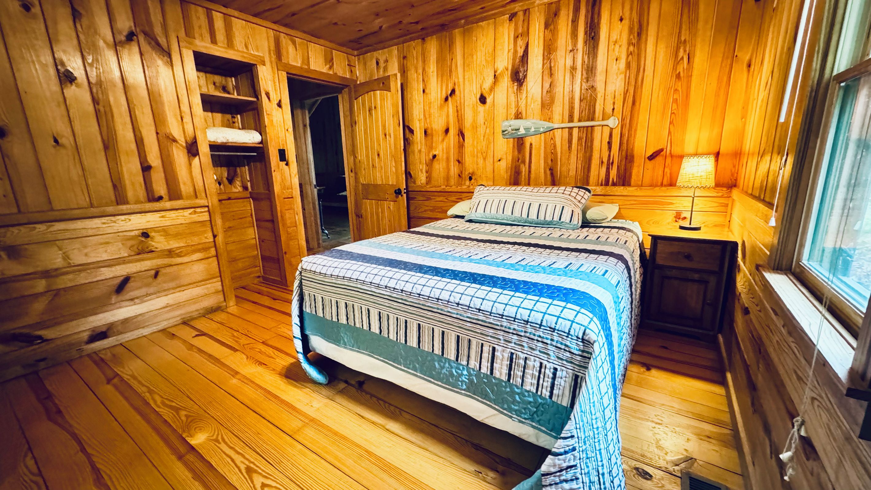A wooden cabin room with a bed, a desk, and a window.