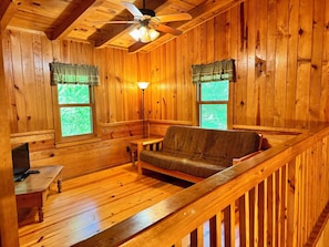 Interior de una cabaña de madera con sofá, mesa, lámpara y ventilador de techo.
