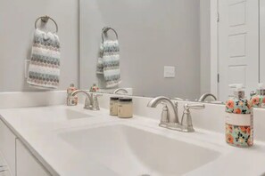 Une salle de bain avec un lavabo blanc, deux robinets et un miroir. Deux serviettes sont accrochées à des crochets, et un distributeur de savon à motifs floraux est posé sur le lavabo.