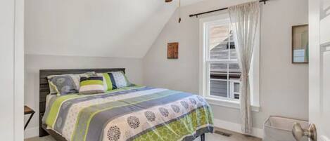 Ein Schlafzimmer mit einem Bett, einem Deckenventilator, einem Fenster mit Vorhängen und einem Wäschekorb.