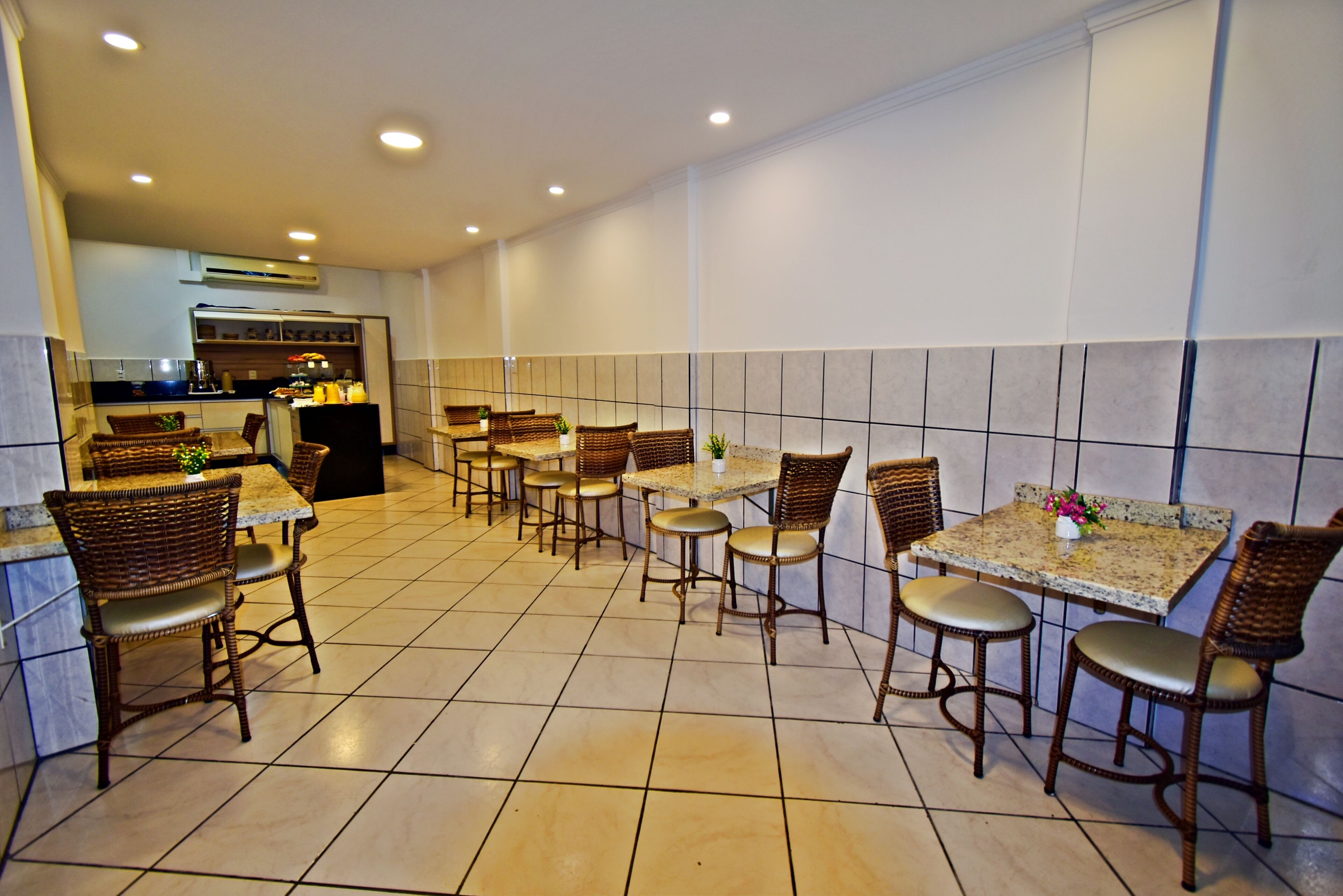 A restaurant with multiple tables and wicker chairs, a kitchen in the background, and tiled walls.