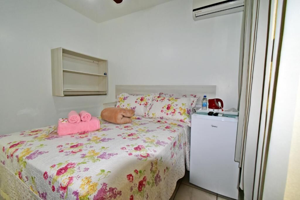 A bedroom with a bed, a small white refrigerator, and a wall-mounted shelf.