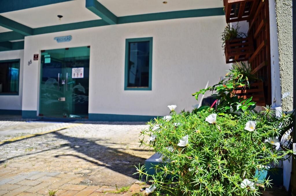 Un edificio con fachada blanca y detalles en color verde azulado, una entrada con puerta de vidrio y una ventana pequeña. En primer plano, hay una planta en maceta con flores blancas.
