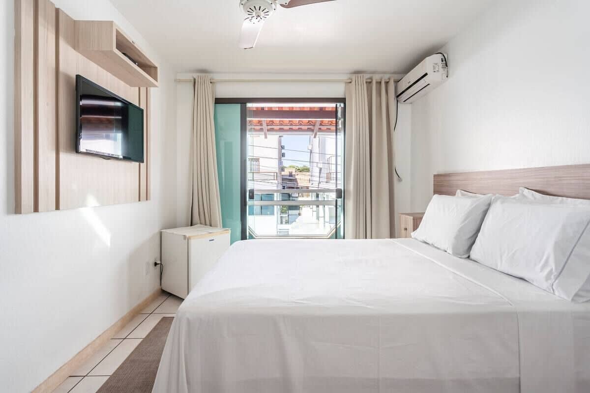 A hotel room with a bed, a TV mounted on the wall, a window with curtains, and an air conditioning unit.