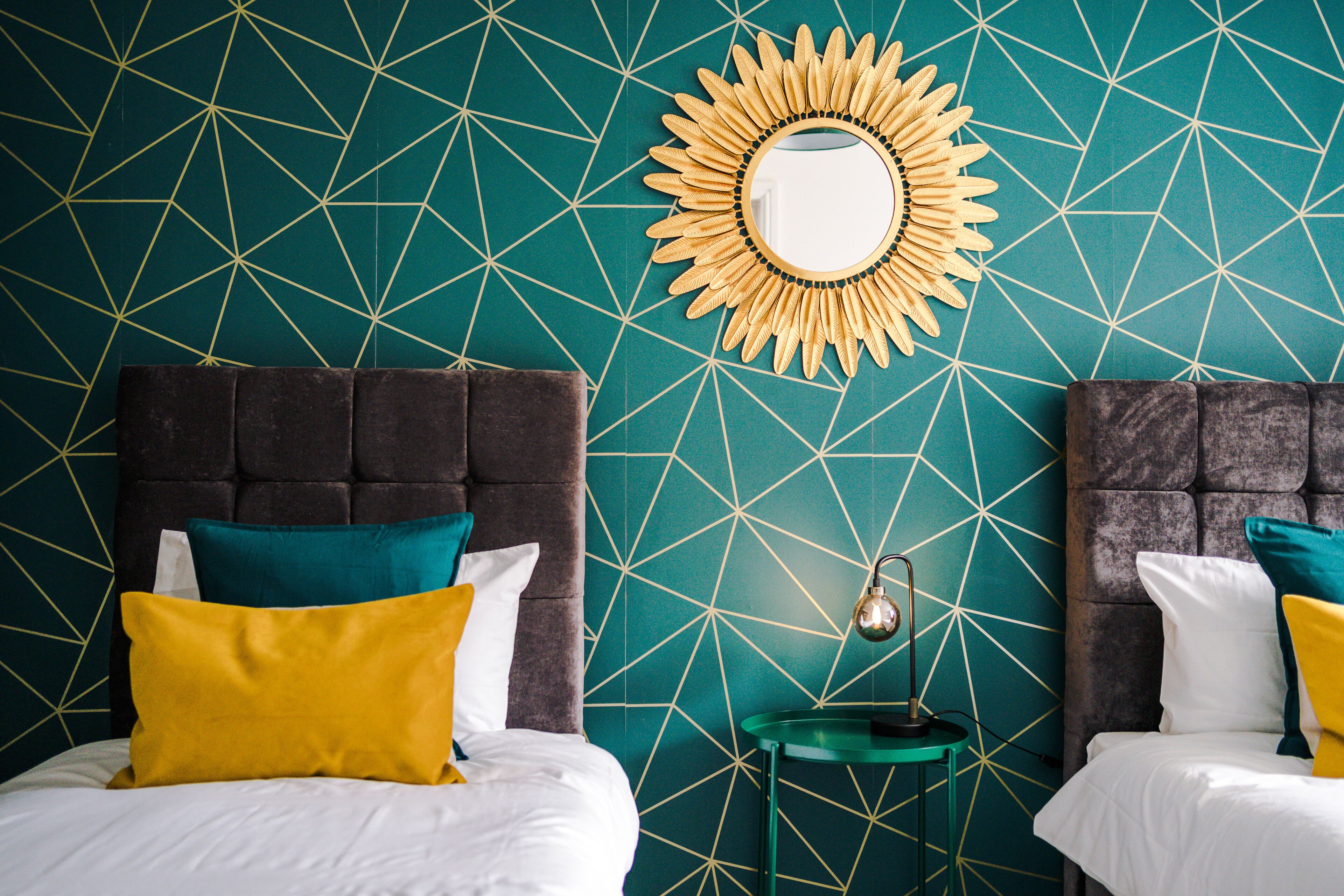 A room with teal patterned wallpaper, a sunburst mirror, two beds with dark headboards, and a small table with a lamp.