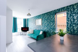 A modern living room with a teal sofa, a flat-screen TV, a dining table, and a potted plant.