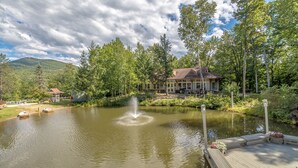 A serene lakeside setting with a wooden dock, a fountain, and a house surrounded by lush greenery. - Nordic Village Townhouse, Storyland, Mountains, Pools (Bartlett)