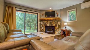 A living room with a stone fireplace, leather sofas, a flat-screen TV, and a large window with curtains. - Nordic Village Townhouse, Storyland, Mountains, Pools (Bartlett)
